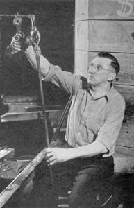 Applying the Stem: Here the gaffer, seated at his bench and using parrot shears, is steadying a gather of glass, as it is applied to the bulb with a pontil rod that will form the goblet stem.