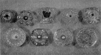 Eight Designs of American Cast Knobs: All are of clear glass. Top row, left to right: Ribbed mushroom design; hollow rosette type and broken knob of same design showing hollow center; swirled mushroom; and design imitating cut glass. Lower row, left to right: Design of fine diamond shapes surrounding deep central cup; hollow crown-shaped rosette; swirl ribs on flat face and conventionalized floral design, also found on Sandwich glass tiebacks.