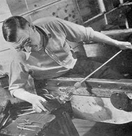 Forming the Foot: A second gather applied with the pontil has provided the material for the goblet foot. Here the gaffer, rotating his blow pipe back and forth on his bench arms, is shaping the foot with a palette of applewood. To prevent burning it is frequently dipped in water as the shaping progresses.