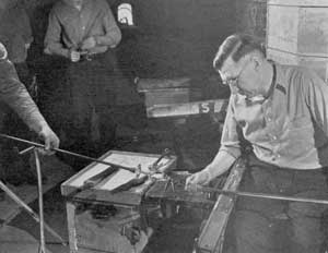 Freed from the Blow Pipe: Here the goblet, firmly attached to the pontil rod, has just been removed by the gaffer from the lower end of the blow pipe by a sharp blow preparatory to forming the upper part or bowl.