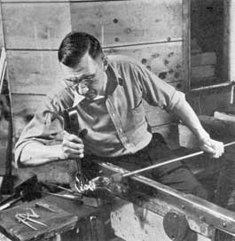 Shaping the Stem: Using a steel fork, as he rolls his blow pipe back and forth on the arms of his bench, the gaffer is giving the goblet stem the desired form. The upper part of the goblet is still attached to the blow pipe and the stem design can be seen.