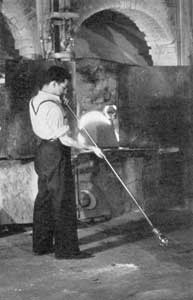 The Start, Blowing the Bulb: Standing before the furnace, a glass blower has expanded the gather of molten glass on his blow pipe. When finished this will be the upper part of the goblet.